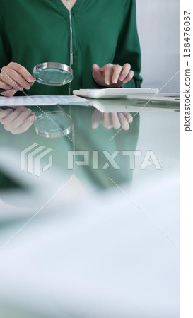 Businesswoman in green blouse carefully reviewing financial reports with magnifying glass and calculator, conducting audit and analyzing data in office. Taxes, audit in business 138476037