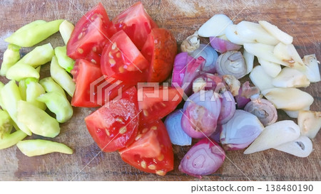 A vibrant assortment of fresh, raw cooking ingredients including chopped tomatoes, shallots, garlic cloves, and green chilies, neatly arranged on a wooden cutting board, ready for meal preparation. 138480190