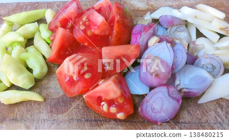 A vibrant display of fresh, chopped vegetables including red tomatoes, purple shallots, white garlic, and green chilies, neatly arranged on a rustic wooden cutting board, ready for cooking a healthy 138480215