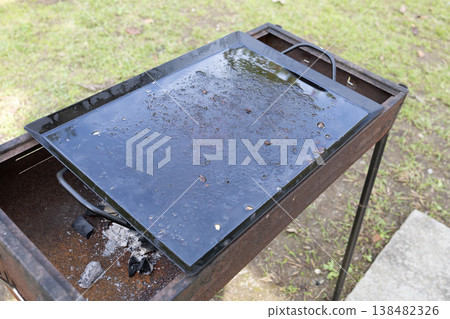 A BBQ grill set up on the lawn in the garden, and a griddle with grease still on it after use. A BBQ grill set up on the lawn in the garden, and a griddle with grease still on it after use. 138482326
