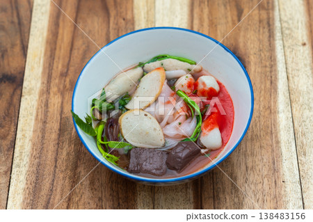 Thai noodle soup with fish balls herbs and vegetables in clear broth served in bowl on wooden table traditional street food fresh and appetizing meal Thai noodle soup with fish balls herbs and vegetables in clear broth served in bowl on wooden table traditional street food fresh and appetizing meal 138483156