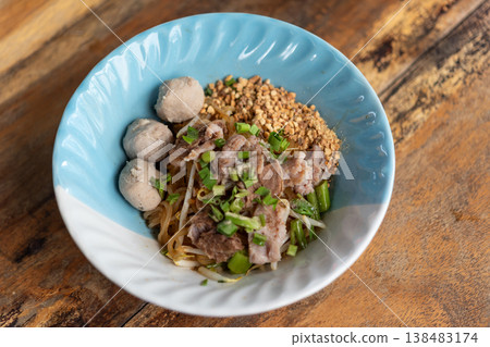 Thai dry noodle dish with pork slices meatballs bean sprouts peanuts and greens in bowl on wooden table authentic street food cuisine savory and flavorful meal 138483174