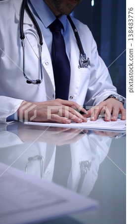Medical professional wearing a white lab coat. Stethoscope. And tie. Sitting at a glass desk. Calculating paperwork. A laptop visible on the side Medical professional wearing a white lab coat. Stethoscope. And tie. Sitting at a glass desk. Calculating paperwork. A laptop visible on the side 138483776