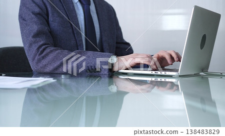 Businessman in blue jacket is focused on typing on laptop at glass desk in modern office, showcasing productivity and concentration, closeup view. Business people concept Businessman in blue jacket is focused on typing on laptop at glass desk in modern office, showcasing productivity and concentration, closeup view. Business people concept 138483829