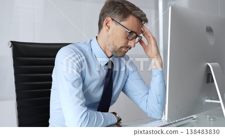 Businessman sitting at a desk in his office, suffering from a severe headache and feeling stressed, tired, and burned out after a long day of intensive work in front of a computer screen Businessman sitting at a desk in his office, suffering from a severe headache and feeling stressed, tired, and burned out after a long day of intensive work in front of a computer screen 138483830