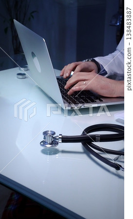 Stethoscope laying on a white reflective desk while medical professional working in clinic office. Typing on laptop and writing notes. Medicine concept 138483887