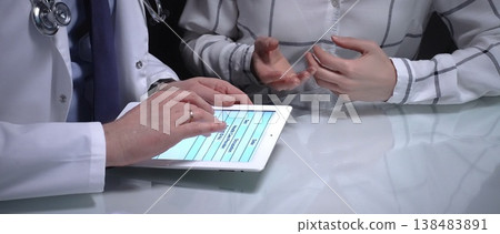 Doctor discussing medical information with a patient using a tablet, showing digital health consultation and modern healthcare communication in a clinic office. Overwork in medicine Doctor discussing medical information with a patient using a tablet, showing digital health consultation and modern healthcare communication in a clinic office. Overwork in medicine 138483891