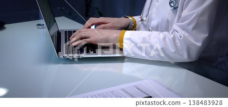 Medical professional wearing a lab coat and stethoscope, typing on a modern laptop computer, working on digital patient records and healthcare data in a clinic or office setting 138483928