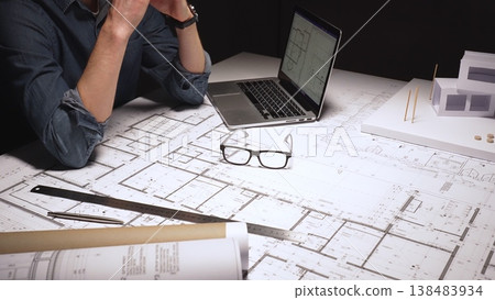 Architect feeling frustrated and stressed, working late on blueprints and building plans, designing a new house with a laptop and architectural model on a desk 138483934