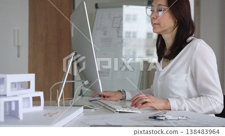 Professional woman architect sitting at office desk, typing on keyboard and looking at monitor, planning building model. Business and architecture concept Professional woman architect sitting at office desk, typing on keyboard and looking at monitor, planning building model. Business and architecture concept 138483964