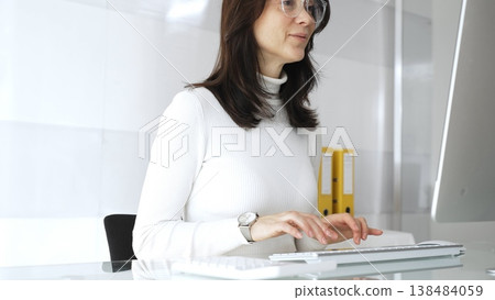 Businesswoman wearing white casual sweater and glasses, typing on a keyboard, performing administrative tasks and navigating digital systems in a modern, clean office environment. Audit in business Businesswoman wearing white casual sweater and glasses, typing on a keyboard, performing administrative tasks and navigating digital systems in a modern, clean office environment. Audit in business 138484059