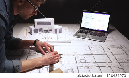 Architect working on new building design project, creating a modern architectural model and drafting technical blueprints with a laptop showing digital plans Architect working on new building design project, creating a modern architectural model and drafting technical blueprints with a laptop showing digital plans 138484070