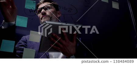 Mature male professional, wearing eyeglasses and suit, working late in a dark office, holding a digital tablet and brainstorming new business strategies with post it notes on a glass board 138484089