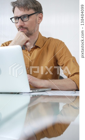 Focused businessman in yellow casual shirt is using laptop and thinking with hand on chin, working on a project in a modern office, vertical portrait. Business people concept 138484099