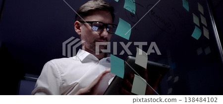Man in eyeglasses organizing thoughts and ideas for business optimization on a clear glass board. Creating a strategic roadmap for innovation and project planning 138484102