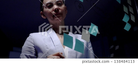 Woman wearing clear frame glasses standing in front of a glass wall covered with sticky notes and drawing a mind map, concentrating on planning a business strategy Woman wearing clear frame glasses standing in front of a glass wall covered with sticky notes and drawing a mind map, concentrating on planning a business strategy 138484113