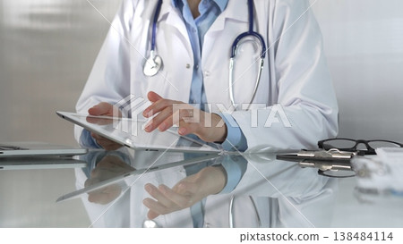 Doctor woman, wearing a blue blouse under a medical coat, is using digital tablet in modern healthcare setting, interacting with patient data, close-up. Medicine concept Doctor woman, wearing a blue blouse under a medical coat, is using digital tablet in modern healthcare setting, interacting with patient data, close-up. Medicine concept 138484114