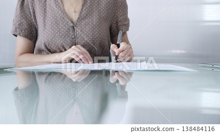 Businesswoman in brawn dotted blouse is signing a contract at a sleek glass desk in a modern office, showcasing professionalism and focus in her work environment, closeup. Business people concept 138484116
