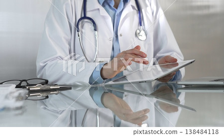 Doctor woman, wearing a blue blouse under a medical coat, is using digital tablet in modern healthcare setting, interacting with patient data, close-up. Medicine concept Doctor woman, wearing a blue blouse under a medical coat, is using digital tablet in modern healthcare setting, interacting with patient data, close-up. Medicine concept 138484118
