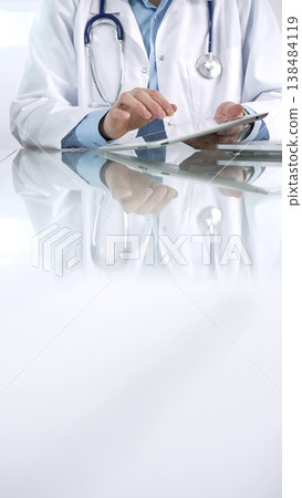 Unrecognizable female doctor in blue blouse, stethoscope and white coat is using digital tablet in modern healthcare setting, interacting with patient data, vertical view. Medicine and science 138484119