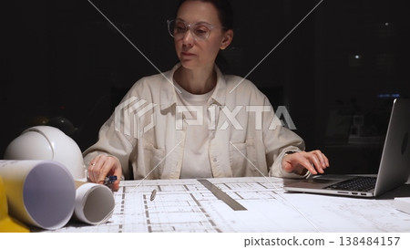 Female architect working late on detailed building blueprints using ruler and pencil, modern engineer designing construction plans at desk with laptop and hardhat 138484157