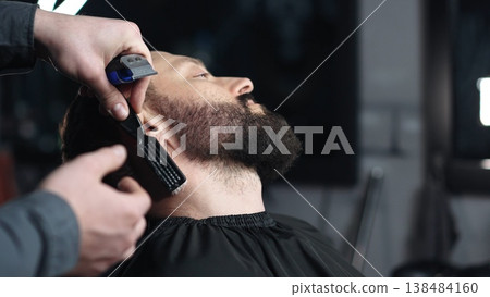 Professional barber trimming client's beard with electric razor, delivering precise styling within contemporary grooming salon setting 138484160