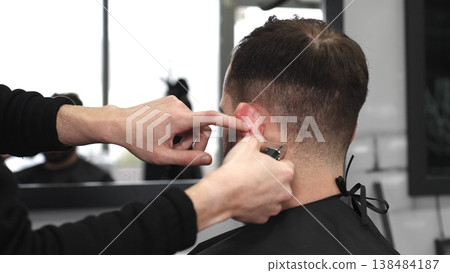 Professional barber trimming male client's hair with electric razor, delivering precise grooming service in contemporary, well lit barbershop setting Professional barber trimming male client's hair with electric razor, delivering precise grooming service in contemporary, well lit barbershop setting 138484187