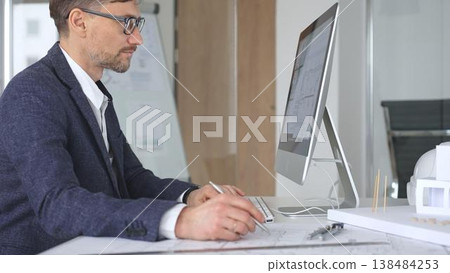 Architect working on computer in design office, planning and developing building projects with blueprints and models. Business and architecture concept Architect working on computer in design office, planning and developing building projects with blueprints and models. Business and architecture concept 138484253