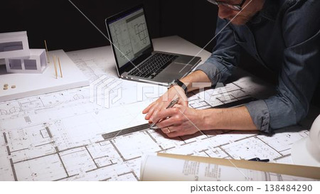 Man architect carefully measuring building blueprints with a compass, working on a new modern house design project, using laptop and scale model in a dimly lit studio Man architect carefully measuring building blueprints with a compass, working on a new modern house design project, using laptop and scale model in a dimly lit studio 138484290