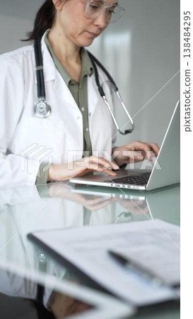 Healthcare female professional in white coat with stethoscope typing on laptop, managing patient records and telehealth data at desk. Medicine, health care and science concept 138484295