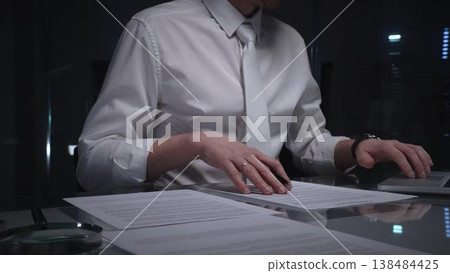 Man in white shirt reviewing legal documents, checking details of a contract with a magnifying glass on a dimly lit desk. Audit, business and overworking concepts 138484425