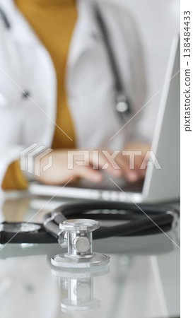 Stethoscope and clipboard with medical documents are on the table near female doctor working on laptop, managing patient data in clinic. Concept of medicine, healthcare and science 138484433