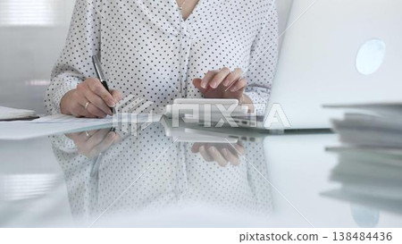 Focused businesswoman at a sleek glass desk in a contemporary office, using a calculator to crunch numbers and jotting down meticulous notes. Taxes, audit in business 138484436