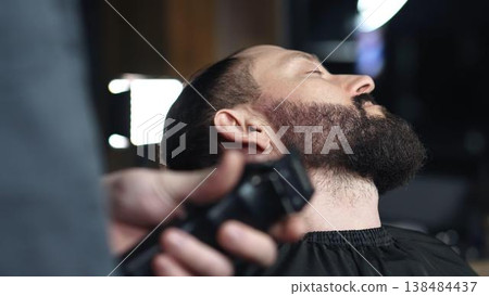 Professional barber trimming client's beard with electric razor, delivering precise styling within contemporary grooming salon setting 138484437