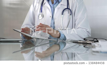 Doctor woman, wearing a blue blouse under a medical coat, is using digital tablet in modern healthcare setting, interacting with patient data, close-up. Medicine concept Doctor woman, wearing a blue blouse under a medical coat, is using digital tablet in modern healthcare setting, interacting with patient data, close-up. Medicine concept 138484438