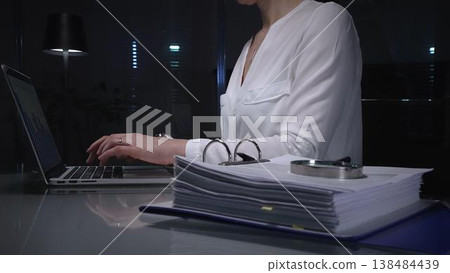 Businesswomen working overtime in a dark office, typing on laptop with financial charts and data reports on desk at night. Audit and taxes in business Businesswomen working overtime in a dark office, typing on laptop with financial charts and data reports on desk at night. Audit and taxes in business 138484439