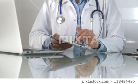 Doctor woman, wearing a blue blouse under a medical coat, is using digital tablet in modern healthcare setting, interacting with patient data, close-up. Medicine concept 138484450