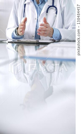 Unknown doctor woman, wearing a stethoscope, blue blouse under a lab coat, is gesturing with hands overs a glass desk with a laptop in clinic office, close up vertical view. Medicine and science Unknown doctor woman, wearing a stethoscope, blue blouse under a lab coat, is gesturing with hands overs a glass desk with a laptop in clinic office, close up vertical view. Medicine and science 138484457