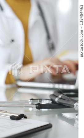 Stethoscope and clipboard with medical documents are on the table near female doctor working on laptop, managing patient data in clinic. Concept of medicine, healthcare and science Stethoscope and clipboard with medical documents are on the table near female doctor working on laptop, managing patient data in clinic. Concept of medicine, healthcare and science 138484462