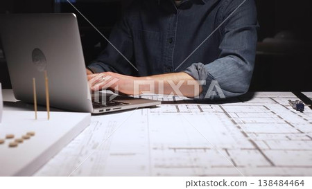 Architect working late at night, typing on a laptop with blueprints and a model on the desk, focusing on architectural design and urban planning in a dimly lit office 138484464