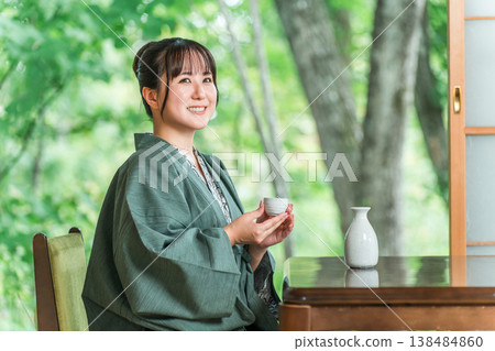 A young Asian woman in a yukata haori relaxing on the veranda of a guest room in a ryokan, drinking sake A young Asian woman in a yukata haori relaxing on the veranda of a guest room in a ryokan, drinking sake 138484860