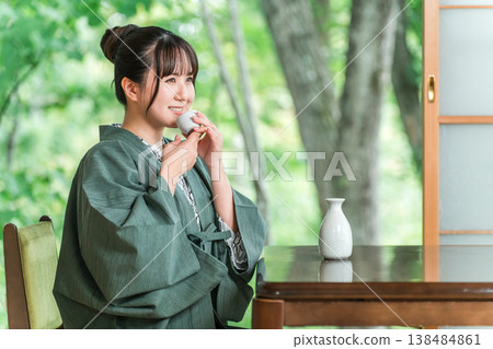一位身穿浴衣的年輕亞洲女子在旅館客房的門廊上放鬆地喝著清酒。 一位身穿浴衣的年輕亞洲女子在旅館客房的門廊上放鬆地喝著清酒。 138484861