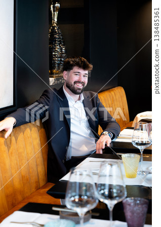 Male waiter in suit leaning on booth, relaxed manager overseeing table settings with glassware, modern artwork backdrop, warm lighting Male waiter in suit leaning on booth, relaxed manager overseeing table settings with glassware, modern artwork backdrop, warm lighting 138485361