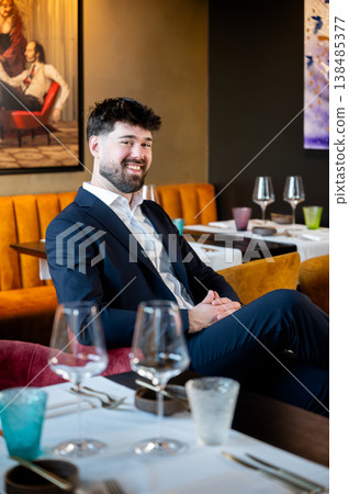 Male waiter in suit smiling candidly at table, authentic dining moment with glassware and warm interior light, approachable service 138485377