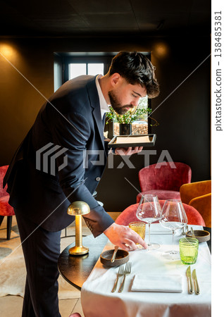 Male waiter setting table with meticulous care, placing glassware and cutlery on crisp linen, small brass lamp casting warm glow, modern Male waiter setting table with meticulous care, placing glassware and cutlery on crisp linen, small brass lamp casting warm glow, modern 138485381