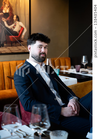 Male waiter in suit seated at upscale restaurant booth, poised with folded hands, set table with wine glasses and folded napkins, warm 138485404