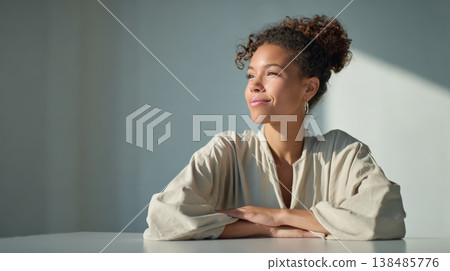 Young businesswoman with curly hair, wearing a beige shirt, sitting at a white table with arms crossed, smiling and looking away, contemplating future opportunities in a sunlit room 138485776