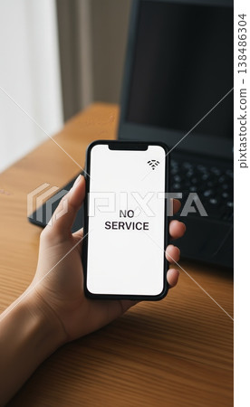 Person holding a smartphone with no service displayed on the screen and a disconnected wifi icon, indicating a connectivity problem while a laptop is in the background 138486304