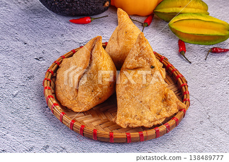 Crispy golden samosas plated with fresh chili, avocado, and star fruit on a textured background. 138489777