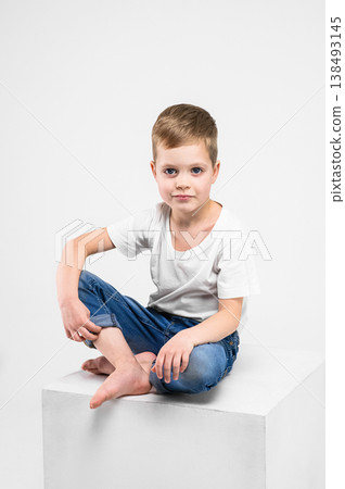 Studio portrait of a cute young boy sitting barefoot on a white cube against a clean white background. Minimalistic lifestyle image representing childhood, calmness and innocence. Studio portrait of a cute young boy sitting barefoot on a white cube against a clean white background. Minimalistic lifestyle image representing childhood, calmness and innocence. 138493145